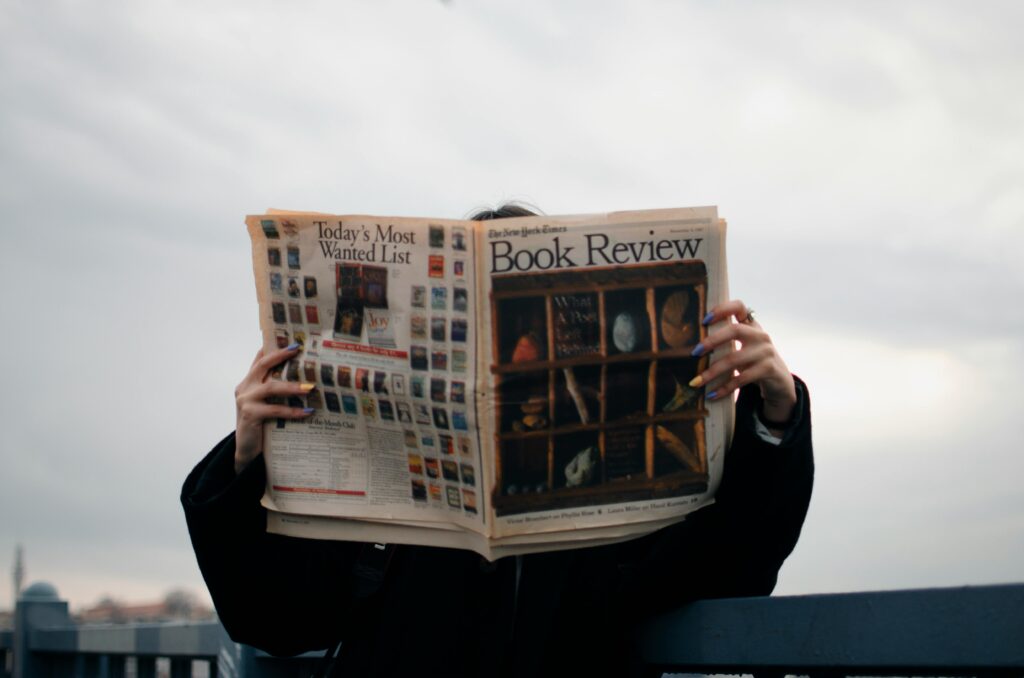 Home Individual holding a book review newspaper with colorful nails against a cloudy sky backdrop.